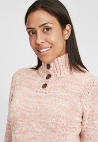 Maglione lavorato a maglia di un morbido rosa con un colletto alto, caratterizzato da tre bottoni marroni sulla patta e dettagli a coste strutturati.