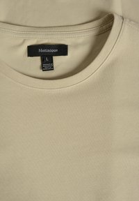 Tricou bej deschis cu guler rotund, realizat dintr-un material neted. Eticheta indică "Matinique," mărimea L, cu instrucțiuni de întreținere pe etichetă.