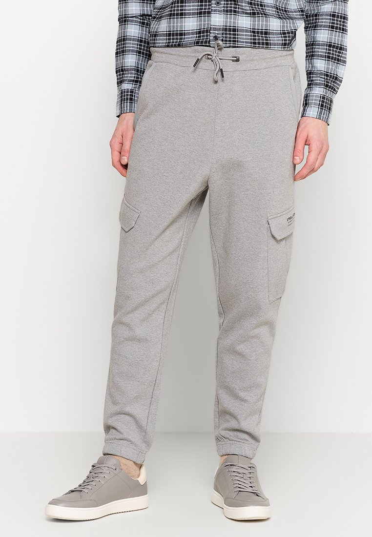 Grijze sweatpants met elastische tailleband en verstelbare trekkoord, voorzien van zijcargo zakken en ribbelmanchetten. Gecombineerd met grijze sneakers.