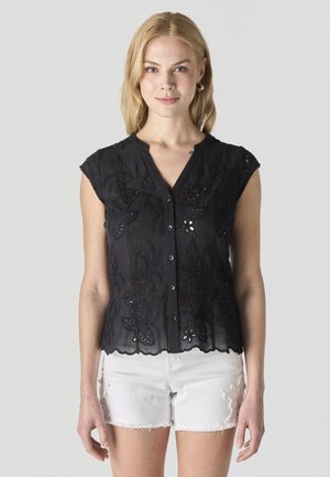 Mujer rubia que lleva una blusa sin mangas negra con botones y bordados, y unos shorts vaqueros blancos con detalles de encaje floral.