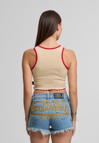 Bežový rebrovaný crop top s červenými lemami a obdĺžnikovým lemom, kombinovaný s modrými denimovými šortkami s žltou vyšívanou aplikáciou "Von Dutch Originals."