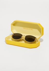 Saulesbrilles dzeltenā oktagona gadījumā. Saulesbrillēm ir tumšas lēcas, zelta krāsas aparatūra un smalkas kājas. Gadījumā iekšpusē ir iespiests "SWAROVSKI".