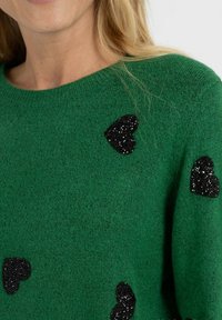 Pull en tricot vert avec un tissu texturé, des manches courtes et des patches en cœur noir pailletés dispersés.