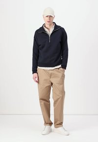 Sudadera de la marina con media cremallera, puños acanalados; sobre una camisa de rayas. Pantalones beige, zapatillas blancas y una gorra beige.