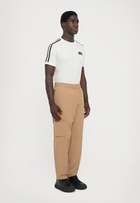 Bijela majica kratkih rukava s crnim prugama na rukavima i logom, uparena s bež cargo hlačama s bočnim džepom i crnim tenisicama.