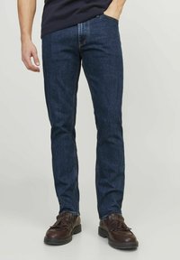 Tumedad denim teksad õhukese lõikega, klassikalise viie tasku disainiga ja kontrastõmblustega, koos pruunide kingadega.