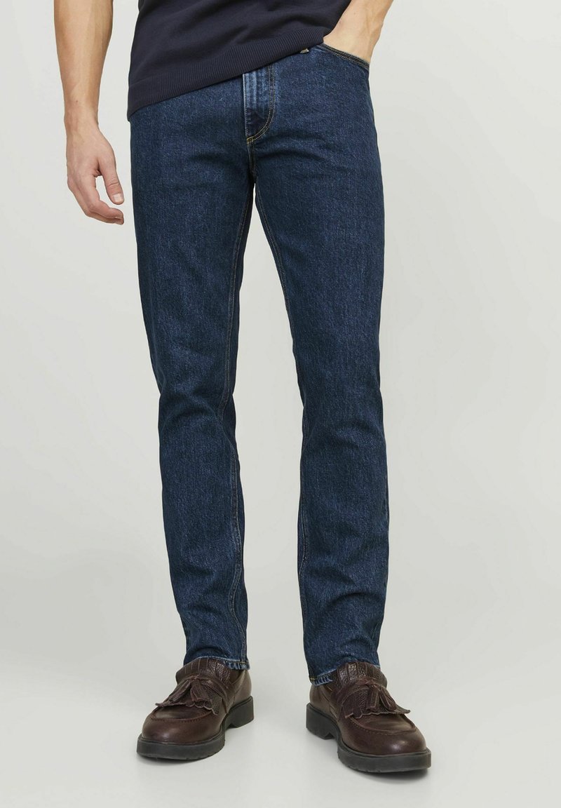 Tumedad denim teksad õhukese lõikega, klassikalise viie tasku disainiga ja kontrastõmblustega, koos pruunide kingadega.