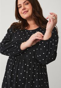 Vestido negro con lunares blancos, con escote redondo y mangas largas y abullonadas. Tejido texturizado, silueta fluida.