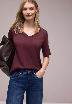 Joven con una camiseta de cuello en V de color vino y jeans azules, llevando un gran bolso de cuero marrón sobre el hombro, con una mano en el bolsillo.