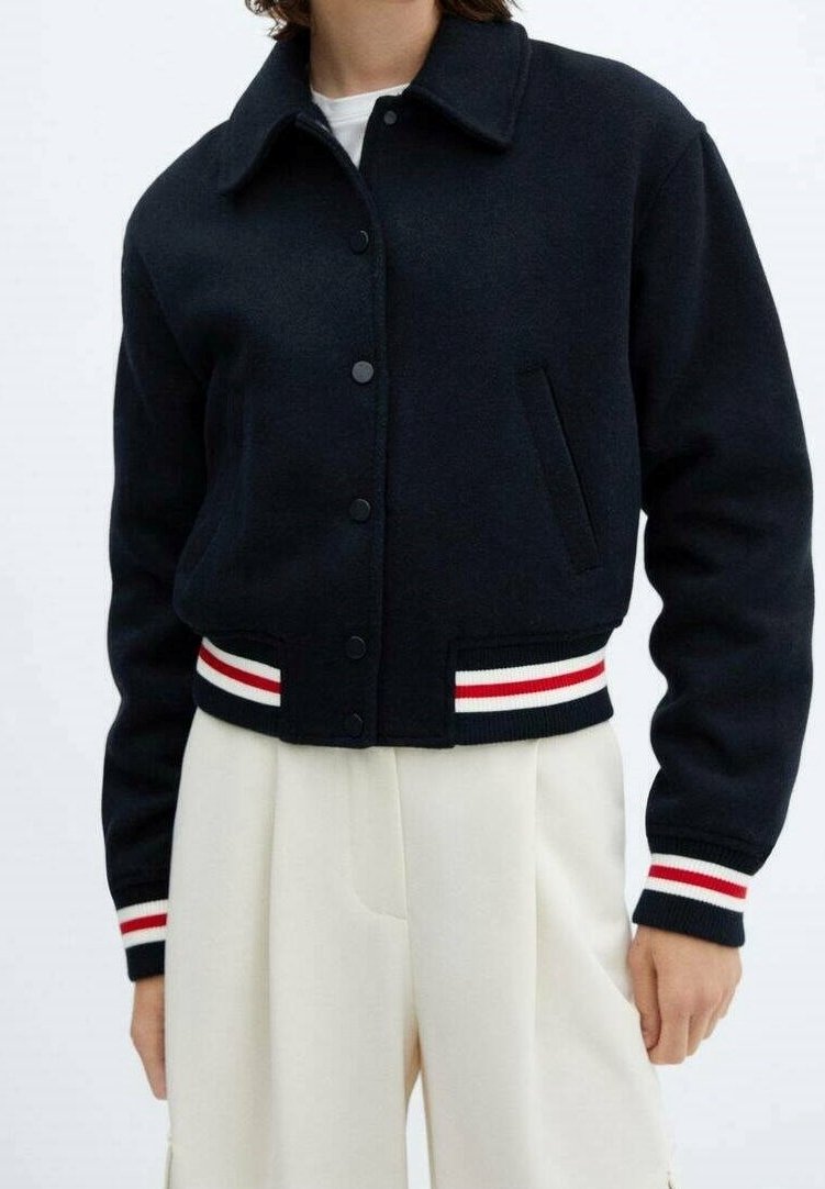 Femme portant une veste noire boutonnée avec des poignets et un ourlet à rayures rouges et blanches, associée à un pantalon crème plissé taille haute.