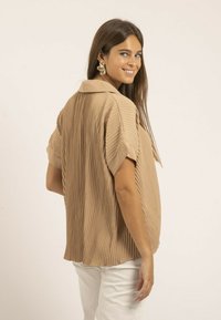 Chemise plissée beige avec un col, des manches courtes et une coupe décontractée. Tissu texturé avec de subtiles lignes verticales et un ourlet arrondi.