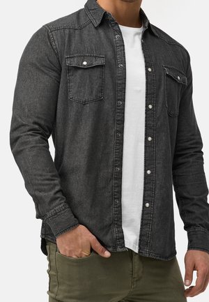 Chemise en denim noir avec deux poches sur la poitrine, boutons-pression, manches longues et col pointu, portée par-dessus un t-shirt blanc.