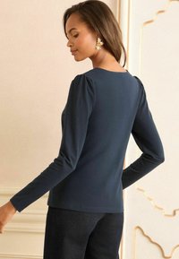 Tumesinise pika varrukaga topp ümarate kaelustega ja kogutud õlavarre detailidega, valmistatud siledast kangast, koos tumedate pükstega.