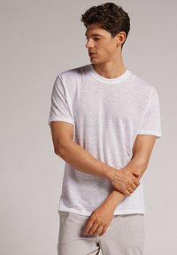 Weißes T-Shirt mit Rundhalsausschnitt aus leichtem, strukturiertem Stoff. Kurze Ärmel und lockere Passform. Das Model posiert mit verschränkten Armen.