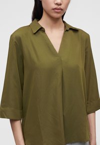 Femme portant un chemisier vert olive à col en V et manches trois-quarts sur un fond clair uni.