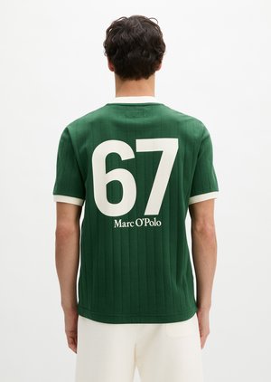 Homme portant un t-shirt vert à manches courtes avec le numéro blanc 67 et le texte « Marc O'Polo » au dos, associé à un short blanc.