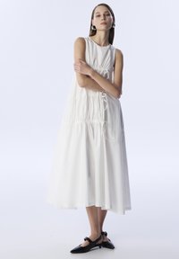 Vestido blanco sin mangas con detalles fruncidos y cordones en la cintura. Combinado con zapatos de vestir negros con hebilla. Textura suave.