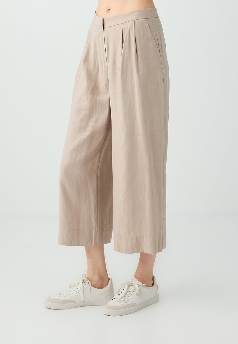 Pantalon beige à jambes larges et raccourci porté avec des baskets blanches basses sur une personne debout devant un fond clair uni.