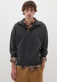 Felpa grigia scura con colletto zip, grande tasca frontale e texture morbida. Sovrapposta a una camicia a quadri marrone.