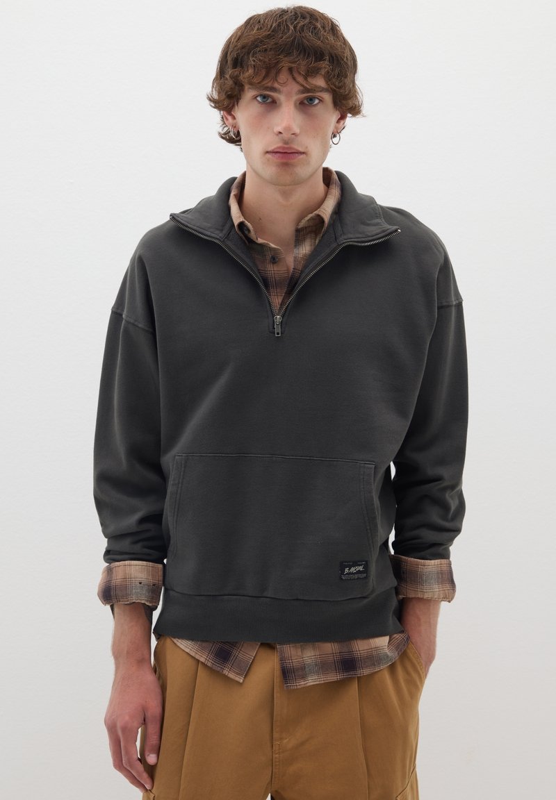 Felpa grigia scura con colletto zip, grande tasca frontale e texture morbida. Sovrapposta a una camicia a quadri marrone.