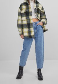 Chaqueta de forro de cuadros en amarillo, blanco, negro y verde, con dos bolsillos frontales. Combinada con unos jeans de tiro alto en azul claro y botines negros.