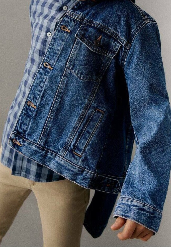 Džínsová bunda v středně modré barvě, se zapínáním na knoflíky, náprsními kapsami a kontrastním prošíváním. Nosená přes modro-bíle kostkovanou košili.