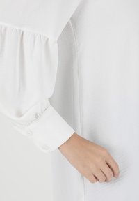 White blouse with gathered long sleeves and buttoned cuffs, featuring a soft, textured fabric and a relaxed fit.
