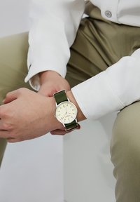 Round watch with a beige dial, silver markers, and a green fabric strap, featuring a minimalist design and a stainless steel case.