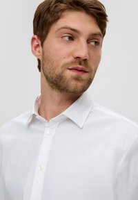 Camicia bianca con colletto, realizzata in un tessuto liscio, dotata di una chiusura davanti con due bottoni visibili e linee semplici e pulite.