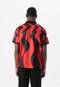 Short-sleeve polo shirt in black with red wavy flame pattern, featuring a black collar and side tag. Smooth, lightweight fabric.