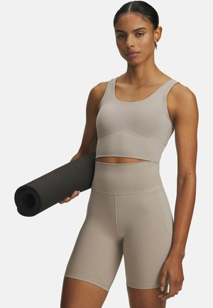 Beige sportbeha en hoog getailleerde korte broek gemaakt van rekbare, gladde stof; heeft een cropped ontwerp en platte naden; zwarte yogamat in één hand gehouden.