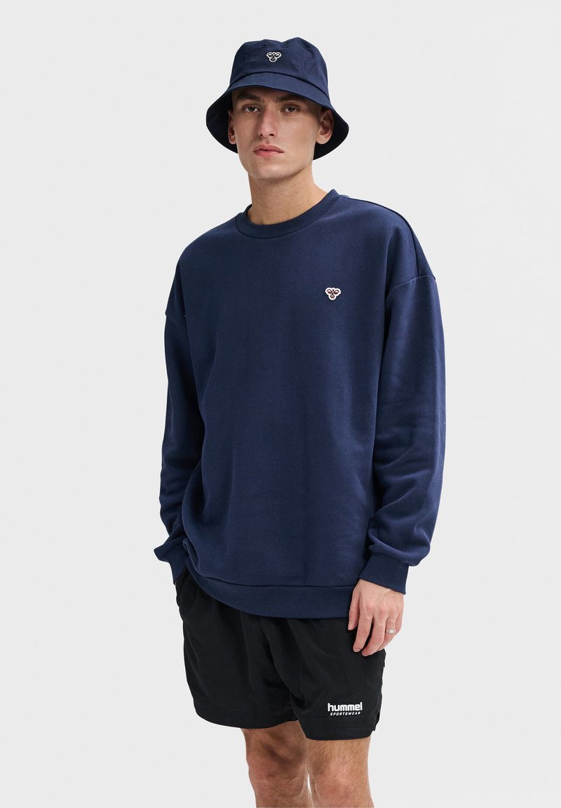 Marineblauwe sweatshirt met een ronde hals en lange mouwen, voorzien van een klein logo op de borst. Gecombineerd met zwarte shorts.