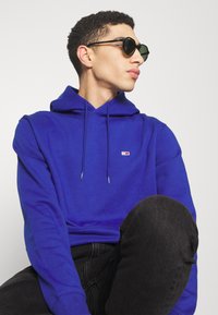 Blauwe hoodie met trekkoorden, met een klein merklogo. G gedragen met zwarte zonnebril en donkere spijkerbroek. Glad stof gevoel.