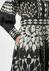 Črno-bela obleka s potiskom, tesno krojena zgornji del, naborki na rokavih in nabrana krila, ki vključujejo geometrijske vzorce in žepe.