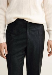 Pantaloni neri a righe sottili realizzati in un tessuto liscio, con un design aderente, chiusura anteriore e dettagli di cucitura discreti. Abbinati a un maglione bianco a coste.