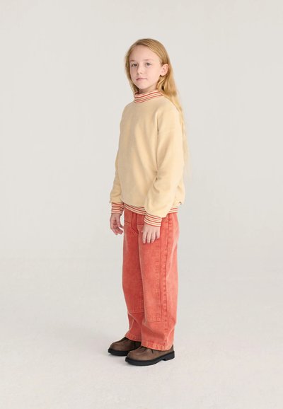 Jeune fille aux longs cheveux blonds portant un pull beige avec des poignets rayés et un pantalon corail large, debout devant un fond clair uni.