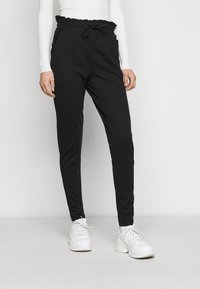 Calças de jogging pretas com uma cintura franzida, cordão ajustável e bolsos laterais. Combinadas com ténis brancos. Textura de tecido suave.