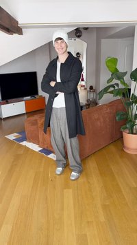 Zwarte jas, wit overhemd, grijze geruite broek en zilveren sneakers. Achtergrond met een bruine bank en planten in een moderne binnenruimte.