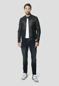 Zwarte leren jas met ritssluitingen op de zakken, gecombineerd met donkerblauwe spijkerbroek met cargozakken, en grijze sportschoenen met witte accenten.