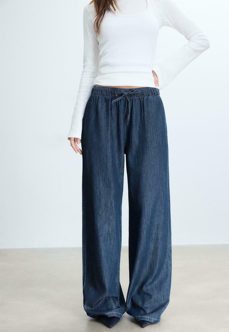 Kobieta ubrana w ciemne jeansy z szerokimi nogawkami i ściągaczem w talii oraz dopasowaną białą bluzkę z długim rękawem, stojąca z jedną ręką na biodrze.