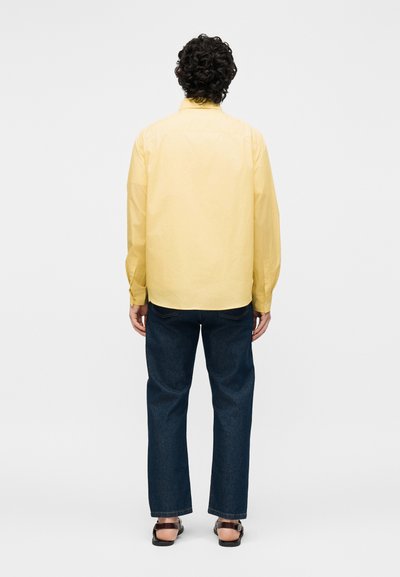 Άτομο με σγουρά μαλλιά που βλέπει προς τα πίσω, φορά κίτρινο μακρυμάνικο πουκάμισο, σκούρα μπλε τζιν και μαύρα παπούτσια χωρίς πτέρνα σε απλό φόντο.