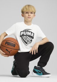 Camiseta blanca con el logo negro de PUMA, pantalones negros y zapatos deportivos negros con acentos en teal. El niño sostiene un balón de baloncesto marrón.