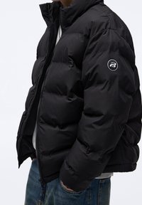 Personne portant une doudoune noire à col montant, logo sur la manche, partiellement dézippée, mains dans les poches d'un jean bleu.