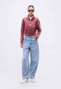 Μπορντό φούτερ με φερμουάρ και μπροστινή τσέπη, σε συνδυασμό με ανοιχτό μπλε τζιν παντελόνι με φαρδιά πόδια και λευκά αθλητικά παπούτσια. Το μοντέλο φοράει μαύρα γυαλιά ηλίου.