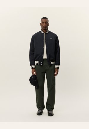 Navy bomber jacket with striped collar, white t-shirt, green trousers, black shoes, and black cap with geometric pattern.