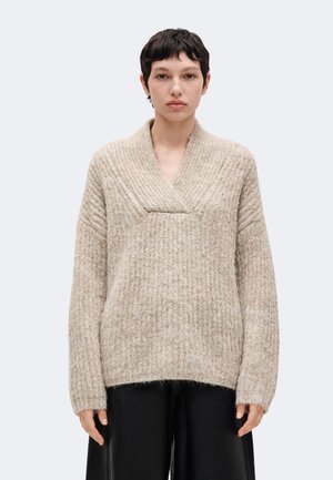Jeune personne aux cheveux courts et foncés portant un pull en maille beige texturé avec un large col en V et un pantalon noir à jambes larges, debout face à l’avant.