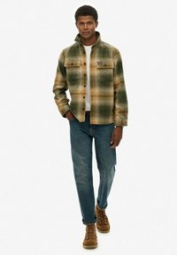 Camicia scozzese verde e beige, con colletto, chiusura con bottoni e due tasche sul petto. Abbinata a jeans blu e stivali marroni.