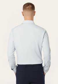 Lichtblauwe blouse met lange mouwen en een knoopsluiting, gladde textuur, rechte snit, en marineblauwe broek. Achteraanzicht toont de knopen op de mouwen en een eenvoudig ontwerp.