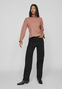 Różowy top z długim rękawem i wysokim kołnierzem, dopasowany do czarnych spodni z wysokim stanem. Modelka nosi czarne botki na obcasie, na szarym tle.