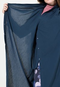 Giacca esterna leggerissima e trasparente di colore blu scuro con bottoni. Il design sovrapposto rivela un capo interno a motivo in tonalità di blu e grigio.